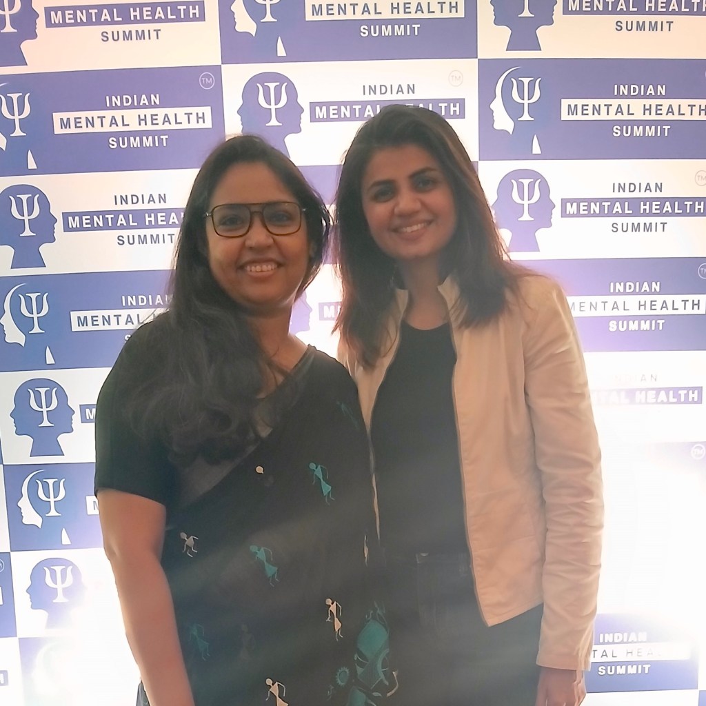 Two female mental health professionals posing in front of the Indian Mental Health Summit (IMHS) backdrop, celebrating collaboration.